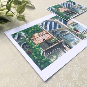 Pu&ograve; includere: Una stampa di un'illustrazione dettagliata di un edificio in stile europeo con un gatto nero. L'edificio ha una porta in legno, finestre e un'insegna con testo giapponese. Lussureggianti piante e fiori verdi circondano l'edificio. Una stampa pi&ugrave; piccola &egrave; sullo sfondo.