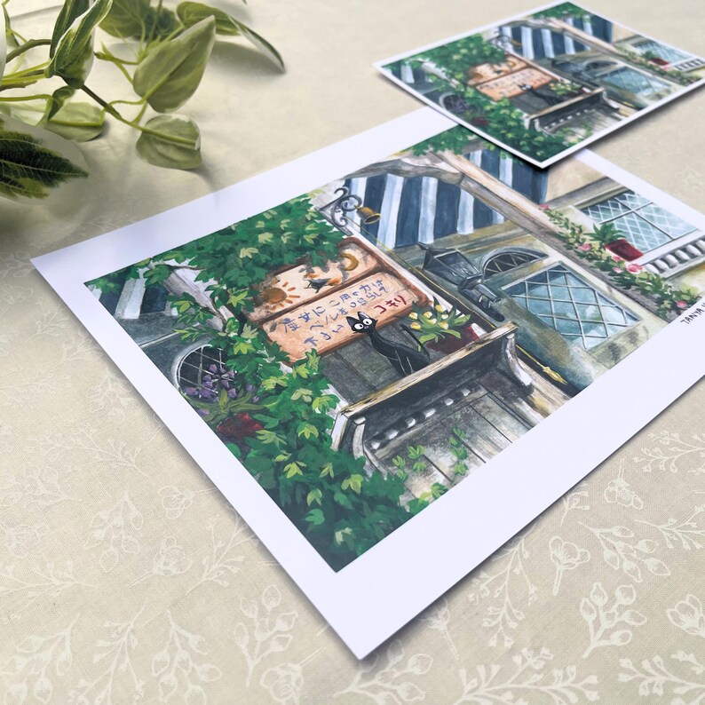 Pu&ograve; includere: Una stampa di un'illustrazione dettagliata di un edificio in stile europeo con un gatto nero. L'edificio ha una porta in legno, finestre e un'insegna con testo giapponese. Lussureggianti piante e fiori verdi circondano l'edificio. Una stampa pi&ugrave; piccola &egrave; sullo sfondo.