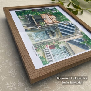 Pu&ograve; includere: Stampa ad acquerello di un edificio con pi&ugrave; finestre, circondato da vegetazione e fiori, incorniciata in una cornice di legno marrone chiaro. La firma dell'artista e l'anno, "Tanya V. 2024", sono visibili.