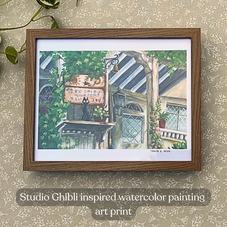 Pu&ograve; includere: Una stampa artistica incorniciata, ispirata a Studio Ghibli. L'opera raffigura un edificio con un'insegna in giapponese, vegetazione lussureggiante e un gatto nero. La cornice &egrave; marrone e la stampa &egrave; firmata "Tanya V. 2014".