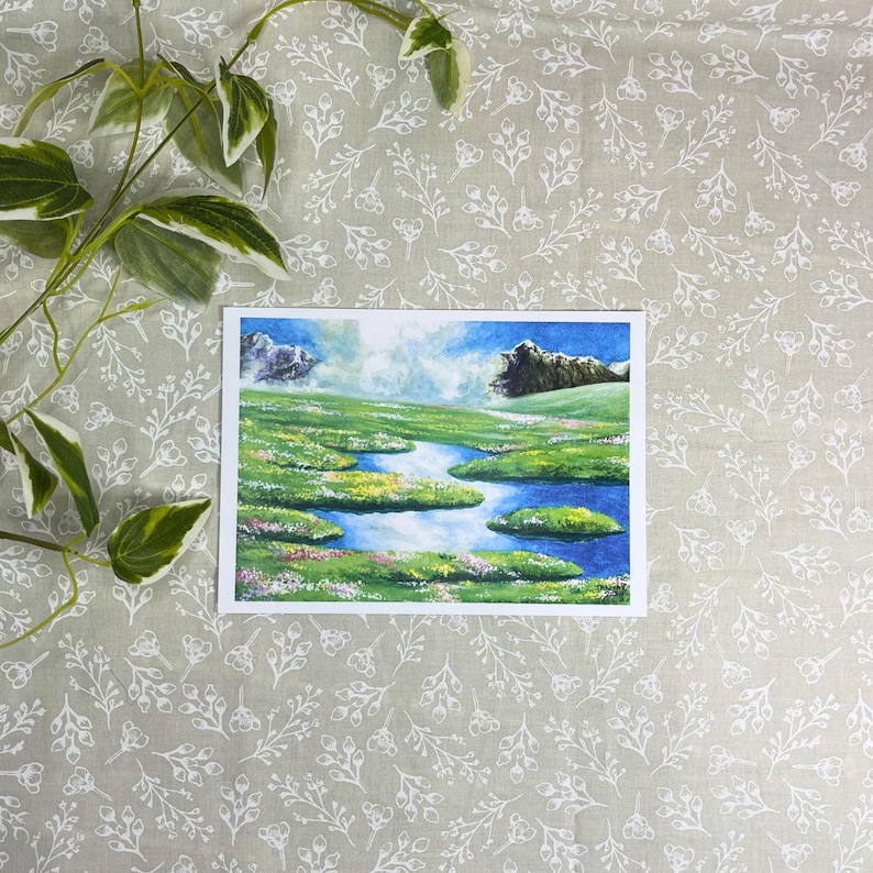 May include: A landscape painting featuring a vibrant green meadow with a winding blue river. The artwork depicts small islands of green and colorful flowers, with mountains in the background under a blue sky with clouds. The painting is on a white rectangular paper.