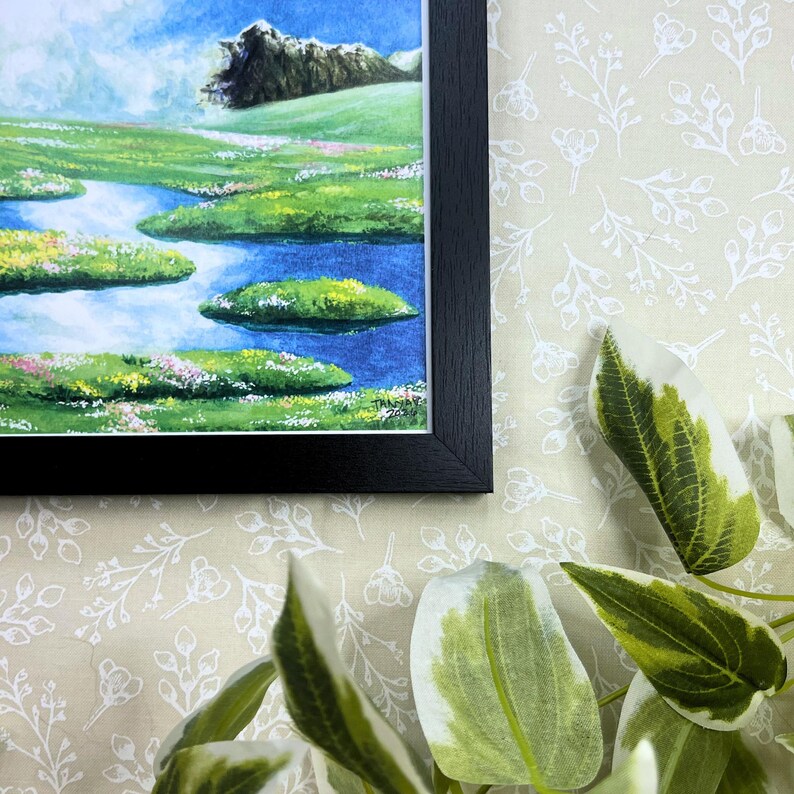 May include: A framed watercolor landscape painting featuring a serene river scene with floating islands of green grass and colorful flowers. The artwork is set against a backdrop of a mountain and a bright blue sky. The frame is black.