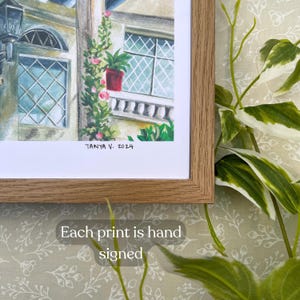 Pu&ograve; includere: Una stampa artistica incorniciata con un acquerello di un edificio con balcone, finestre e piante rampicanti. La stampa &egrave; firmata "Tanya V. 2024". &Egrave; visibile anche il testo "Each print is hand signed".