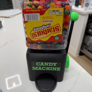May include: A black candy machine with a clear container filled with colorful Starburst jellybeans. The machine has a green handle and the words "CANDY MACHINE" in green letters.