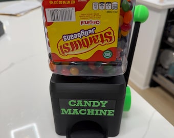 May include: A black candy machine with a green handle and a clear plastic container filled with Starburst jellybeans. The container features a label with the text Starburst Jellybeans Original, net weight 54 oz (1.5 kg), and the base has the text CANDY MACHINE.
