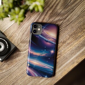 Puede incluir: Una funda de teléfono inteligente con un diseño de galaxia vibrante con nebulosas arremolinadas en tonos de azul, morado y dorado. La funda está sobre una superficie de madera, con una cámara y vegetación en el fondo.