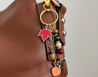May include: A gold-toned keychain with autumn-themed charms, including a red maple leaf, acorn, pumpkin, and a squirrel. The charms are attached to a gold chain and a ring. The keychain is hanging on a brown leather bag.