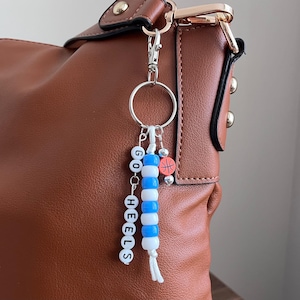 K&ouml;nnte beinhalten: Eine braune Lederhandtasche mit einem silbernen Schl&uuml;sselanh&auml;nger. Der Schl&uuml;sselanh&auml;nger hat wei&szlig;e und blaue Perlen mit der Aufschrift "GO HEELS" und einen kleinen Basketball-Anh&auml;nger. Die Tasche hat goldfarbene Beschl&auml;ge.