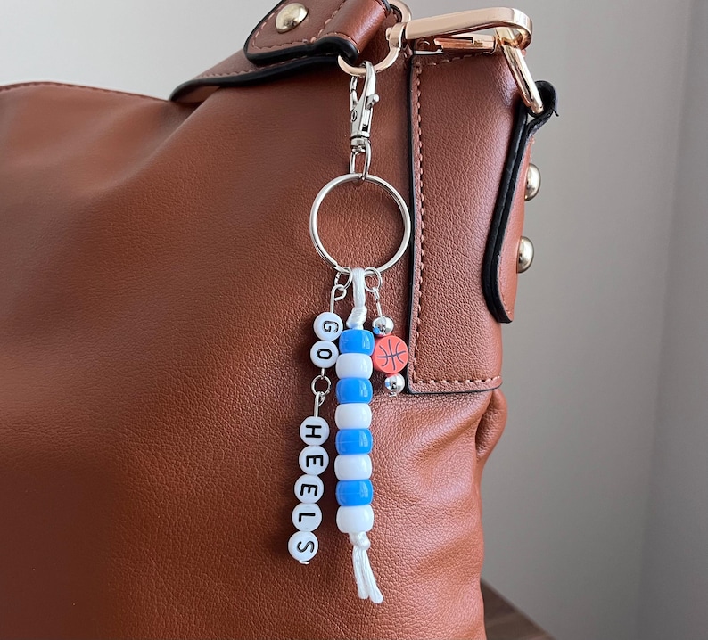 K&ouml;nnte beinhalten: Eine braune Lederhandtasche mit einem silbernen Schl&uuml;sselanh&auml;nger. Der Schl&uuml;sselanh&auml;nger hat wei&szlig;e und blaue Perlen mit der Aufschrift "GO HEELS" und einen kleinen Basketball-Anh&auml;nger. Die Tasche hat goldfarbene Beschl&auml;ge.