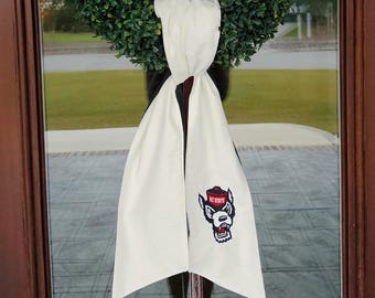 Può includere: Sciarpa da porta color crema con logo ricamato NC State Wolfpack. La sciarpa è drappeggiata su una ghirlanda verde. Il logo presenta una testa di lupo rossa e bianca. Un elemento decorativo per la porta.