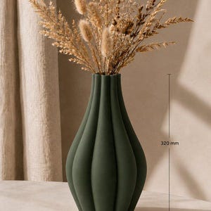 Può includere: Un vaso in ceramica verde scuro opaco con una silhouette bulbosa e scanalata verticalmente. Il vaso contiene un bouquet di fiori ed erbe essiccate color beige. Le dimensioni indicate sono 320 mm di altezza e 175 mm di larghezza.