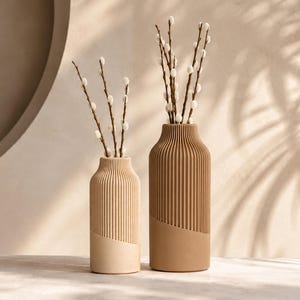 Può includere: Due vasi cilindrici con texture a coste verticali sulle sezioni superiori e basi lisce. Il vaso più corto è beige chiaro, mentre quello più alto è color marrone chiaro. Entrambi contengono rami marroni sottili con piccoli germogli bianchi.