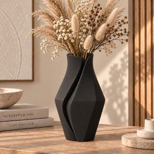 Può includere: Un vaso in ceramica nero opaco con una silhouette geometrica asimmetrica caratterizzata da una profonda fessura verticale. Il vaso è riempito con una composizione di fiori secchi composta da erba della pampa beige, gipsofila bianca e piccoli baccelli marroni.