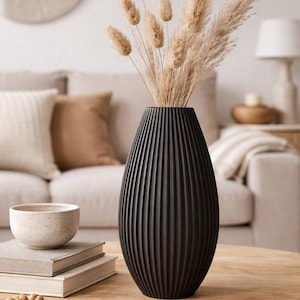 Può includere: Un vaso nero opaco con una texture a coste verticali, contenente un bouquet di erba della pampa e code di lepre essiccate color beige. Il vaso ha una forma ovale affusolata e poggia su una superficie in legno chiaro e liscia.
