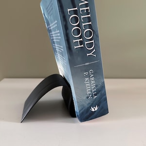 May include: A book titled "Melody Looh" by Gabriella P. Kjeilen, supported by a black, curved book stand. The book has a blue cover with a forest scene. The stand is on a white surface.
