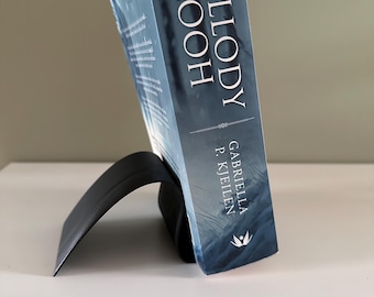 Könnte beinhalten: Ein Buch mit dem Titel "Melody Looh" von Gabriella P. Kjeilen, das von einem schwarzen, gebogenen Buchständer gehalten wird. Das Buch hat einen blauen Einband mit einer Waldszene. Der Ständer steht auf einer weißen Oberfläche.