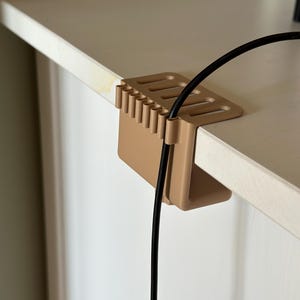Puede incluir: Un organizador de cables de color beige está fijado al borde de una superficie blanca. El organizador tiene una serie de ranuras y un gancho en forma de U para sujetar los cables. Un cable negro está insertado en el organizador.