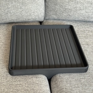 Könnte beinhalten: Ein dunkelgraues rechteckiges Tablett mit gerillter Innenseite. Das Tablett hat ein niedriges Profil und ist so konzipiert, dass es über die Armlehne eines Sofas passt. Das Tablett besteht aus einem matten Material.