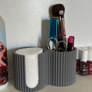 May include: A gray, ribbed, two-section organizer for beauty products. One section holds a stack of white cotton pads, the other contains makeup brushes, scissors, and lipsticks. The organizer sits on a white shelf.