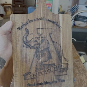 May include: Wooden cutting board with a handle, featuring an engraved elephant graphic and the text "Rather be smokin' in some butt" and "Crimson Tide". The board has a natural wood grain.