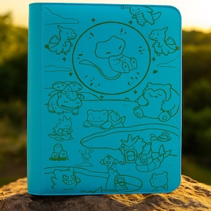 Può includere: Un raccoglitore turchese con un disegno a tratto verde di vari personaggi dei cartoni animati. Il raccoglitore è rettangolare con angoli arrotondati e si trova su una roccia. Lo sfondo è sfocato con un tramonto.