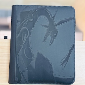 Può includere: Un raccoglitore di carte nero con un'illustrazione in rilievo di una creatura simile a un drago. Il raccoglitore ha una superficie strutturata e un bordo cucito. Il design è elegante e moderno.