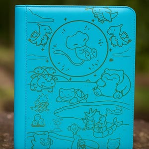 Puede incluir: Una carpeta turquesa con una ilustración dorada de personajes de dibujos animados. La carpeta tiene una textura suave y un borde cosido. Los personajes están dispuestos en un patrón circular, con un personaje central en el medio.