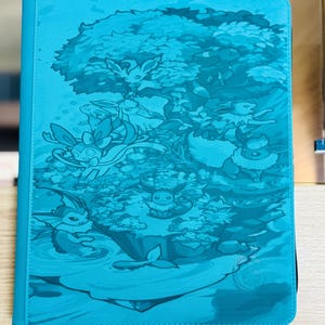 Puede incluir: Una carpeta de color turquesa con una ilustración detallada de personajes de dibujos animados dentro de un diseño de árbol y paisaje. La carpeta tiene un cierre de cremallera y un borde cosido. La obra de arte está en tonos turquesa y azul.