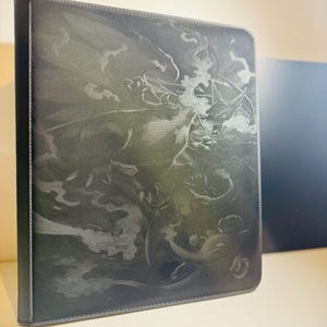 Può includere: Raccoglitore nero con un'illustrazione dettagliata di un drago e altre creature fantasy. La copertina ha un aspetto strutturato e un bordo cucito. L'opera d'arte è in tonalità di grigio e nero, creando un effetto drammatico.