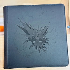 Può includere: Un raccoglitore nero con un'illustrazione stilizzata di Zapdos, un Pokémon uccello leggendario. Il raccoglitore ha una superficie strutturata e un bordo cucito. Il design di Zapdos è in una tonalità più scura di nero.