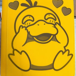 Può includere: Raccoglitore giallo con un personaggio di anatra dei cartoni animati sorridente. L'anatra è delineata in marrone, con cuori marroni sopra. Il raccoglitore ha un bordo cucito.