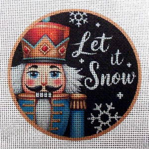 May include: Round Christmas ornament featuring a nutcracker soldier with a red and gold crown, blue jacket, and white hair. The words "Let it Snow" are written in white script on a black background, with snowflakes.