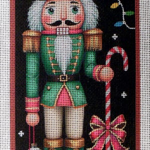May include: A Christmas-themed needlepoint design of a nutcracker soldier. The soldier wears a green uniform with gold accents, a black mustache, and holds a candy cane and ornaments. A string of colorful lights and a red bow complete the festive scene.