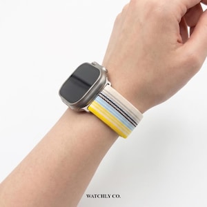 May include: A silver smartwatch with a colorful striped band. The band features yellow, blue, and black stripes against a cream background. The text "WATCHLY CO." is visible at the bottom.