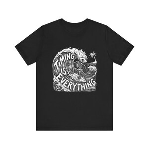 Puede incluir: Camiseta negra con un gráfico blanco de un esqueleto surfeando una ola. El texto "TIMING IS EVERYTHING" está integrado en el diseño de la ola, con una palmera en el fondo. La camiseta está hecha de un material suave.