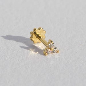 Op de afbeelding: Een gouden oorbel met drie kleine, heldere edelstenen in een cluster. De oorbelpen heeft een bloemvormige achterkant. De oorbel staat tegen een witte achtergrond, met een schaduw naar links.