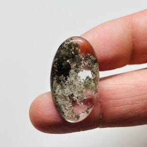 May include: Oval-shaped gemstone with a clear, transparent base. The stone features inclusions of black, green, and white minerals, creating a unique, natural pattern. The gemstone is held between fingers.