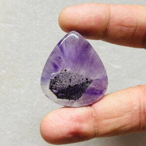 Puede incluir: Una gema en forma de lágrima con un tono púrpura translúcido, que presenta una inclusión oscura y moteada cerca de la base. La piedra se sostiene entre los dedos, mostrando su superficie pulida y sus patrones naturales. El color de la gema varía de púrpura claro a oscuro.