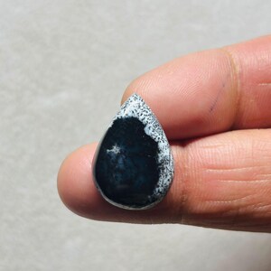 Puede incluir: Una piedra preciosa en forma de lágrima con una superficie pulida. La piedra presenta un centro azul oscuro con bordes moteados blancos. La piedra preciosa se sostiene entre dos dedos, mostrando su tamaño y forma. El patrón y el color únicos de la piedra la convierten en una pieza llamativa.