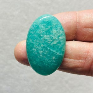May include: A polished, oval gemstone in teal and turquoise hues. The stone has a smooth, glossy surface with subtle internal patterns and a slight sheen. The gemstone is held between fingers.