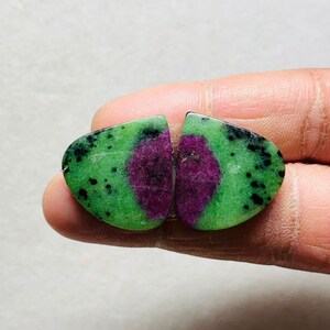 Może przedstawiać: Dwa polerowane kamienie szlachetne zoisytu. Każdy kamień ma żywą zieloną podstawę z czarnymi plamkami i centralnym, głęboko purpurowym rdzeniem. Kamienie są pocięte w zaokrąglony kształt.