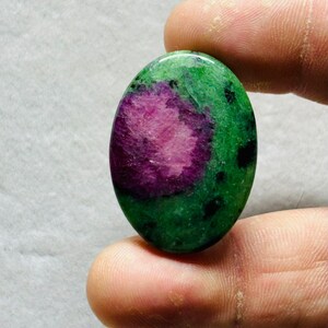 Puede incluir: Gema ovalada con un patrón de color vibrante. La piedra presenta un área central de color magenta intenso, rodeada por un campo verde moteado. Pequeñas inclusiones negras están dispersas por el verde. La gema se sostiene entre los dedos.