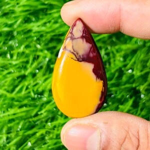 Puede incluir: Una piedra preciosa en forma de lágrima con una paleta de colores vibrantes. La piedra presenta una llamativa combinación de tonos amarillos, burdeos y crema. La piedra preciosa se sostiene entre dos dedos, mostrando su superficie pulida y sus patrones de color únicos.