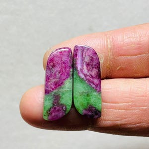 May include: Two polished gemstones, each with a rounded top and a rectangular base. The stones display a vibrant mix of colors, with a deep magenta hue at the top transitioning into a bright green at the bottom. The stones are held between fingers.