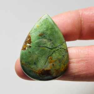 May include: A polished, teardrop-shaped gemstone with a predominantly green hue, accented by brown and black veining and patches. The stone is held between fingers, showcasing its smooth, reflective surface. This natural stone is ideal for jewelry making.