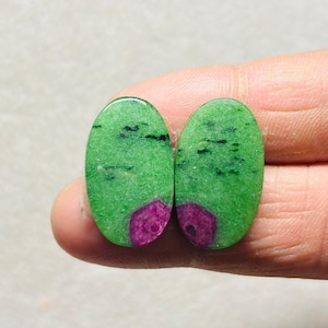 Può includere: Due pietre preziose ovali con una vibrante base verde, punteggiate da inclusioni nere. Ogni pietra presenta una sezione di colore magenta intenso a un'estremità. Le pietre preziose sono tenute tra le dita.