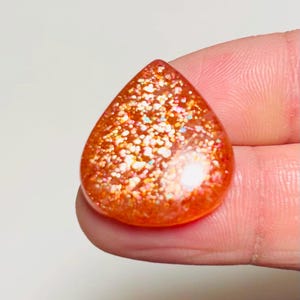 May include: A teardrop-shaped sunstone cabochon featuring a vibrant orange base colour with dense, sparkling metallic inclusions that create a reflective, glittery effect throughout the surface of the polished gemstone.
