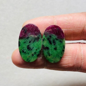 Könnte beinhalten: Zwei ovale Edelsteine. Jeder Stein hat eine tief burgunderfarbene obere Hälfte und eine leuchtend grüne untere Hälfte mit schwarzen Sprenkeln. Die Steine werden zwischen Fingern gehalten, wodurch ihre polierten Oberflächen und natürlichen Muster hervorgehoben werden.