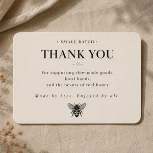 Puede incluir: Una tarjeta beige con esquinas redondeadas, con las palabras "THANK YOU" en una fuente clásica. La tarjeta incluye texto sobre el apoyo a los productos de fabricación lenta y la miel real, junto con una ilustración de abeja. La tarjeta está sobre un fondo de tela neutra.