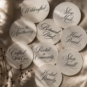 May include: A selection of circular, cream-coloured labels with stylish black calligraphy. The labels display phrases such as "Wildcrafted," "Slow Made," "Botanical Blend," and "Herbal Remedy." They are arranged on a beige fabric.